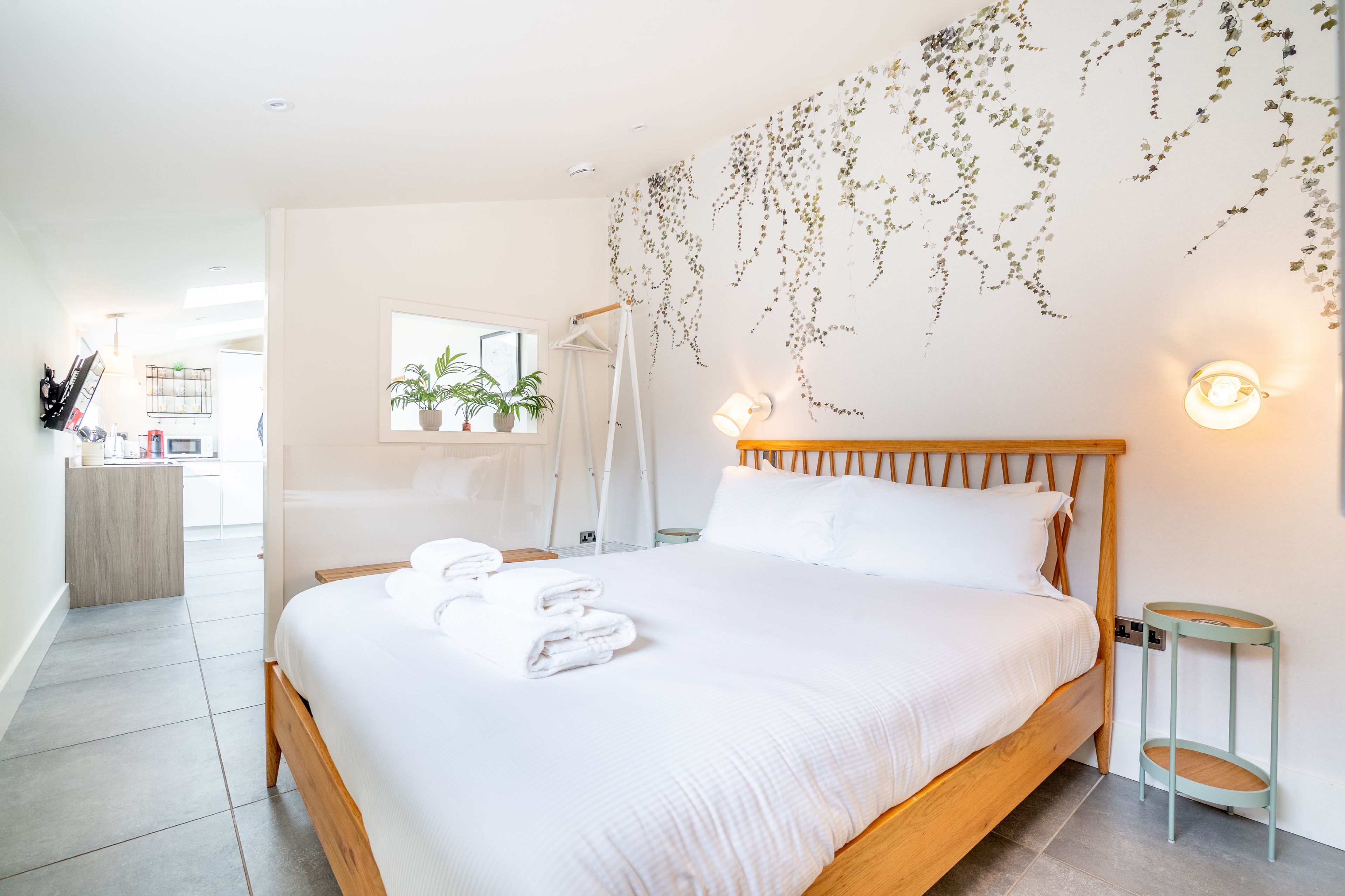 A hotel room with a bed, a bedside table, a wall-mounted light, and a view of the bathroom.
