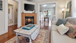 A living room with a fireplace, a flat-screen TV mounted above it, a coffee table with magazines, and a sofa with cushions.