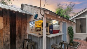 A rustic bar with a wooden exterior, a neon sign for 'Mebbest Lounge', and a Texas flag.