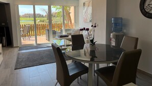 Un coin repas comprenant une table ronde en verre, quatre chaises, un distributeur d’eau et une vue sur une terrasse agrémentée d’une clôture en bois et de verdure.