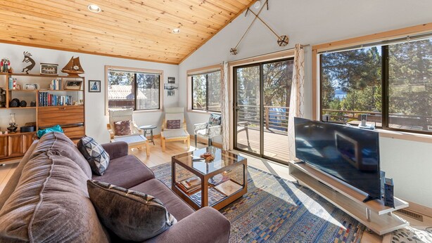 A spacious living room with a wooden ceiling, a large flat-screen TV on a stand, a sofa, a coffee table, and a bookshelf.