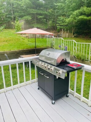 A barbecue grill on a deck with a white railing, a grassy yard, and a wooden fence in the background.