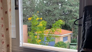 Une fenêtre donnant sur un bâtiment avec un toit rouge, entouré d’arbres et sur lequel on aperçoit une poubelle verte.
