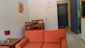 A living room with a sofa, a wooden table with chairs, a red lamp, and a door in the background. - Superstar free Internet free welcome pk (Paphos)