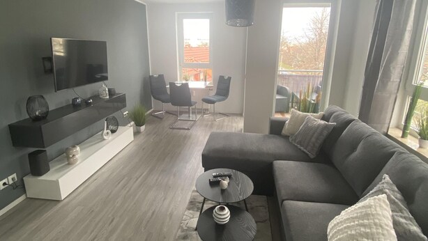 Un salon moderne avec un canapé gris, une table basse et un téléviseur posé sur un meuble blanc. Deux fenêtres laissent entrer la lumière naturelle et offrent une vue sur l’extérieur.