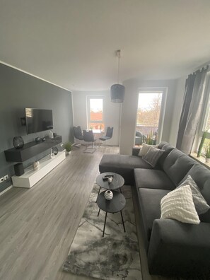 Een moderne woonkamer met een grijze bank, een salontafel en een televisie op een witte console. Twee ramen zorgen voor natuurlijk licht en uitzicht.