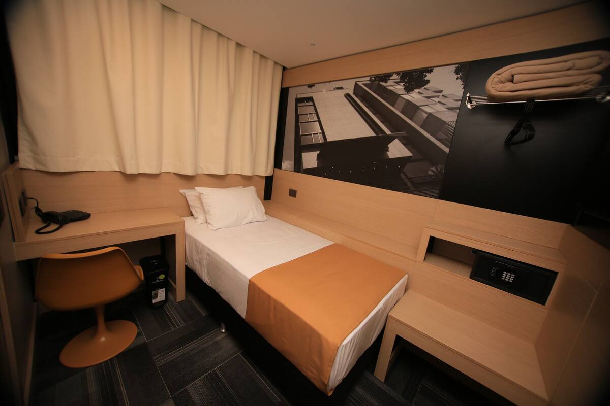 A compact hotel room with a single bed, a desk, a chair, a small table, a wall-mounted TV, and a towel rack.