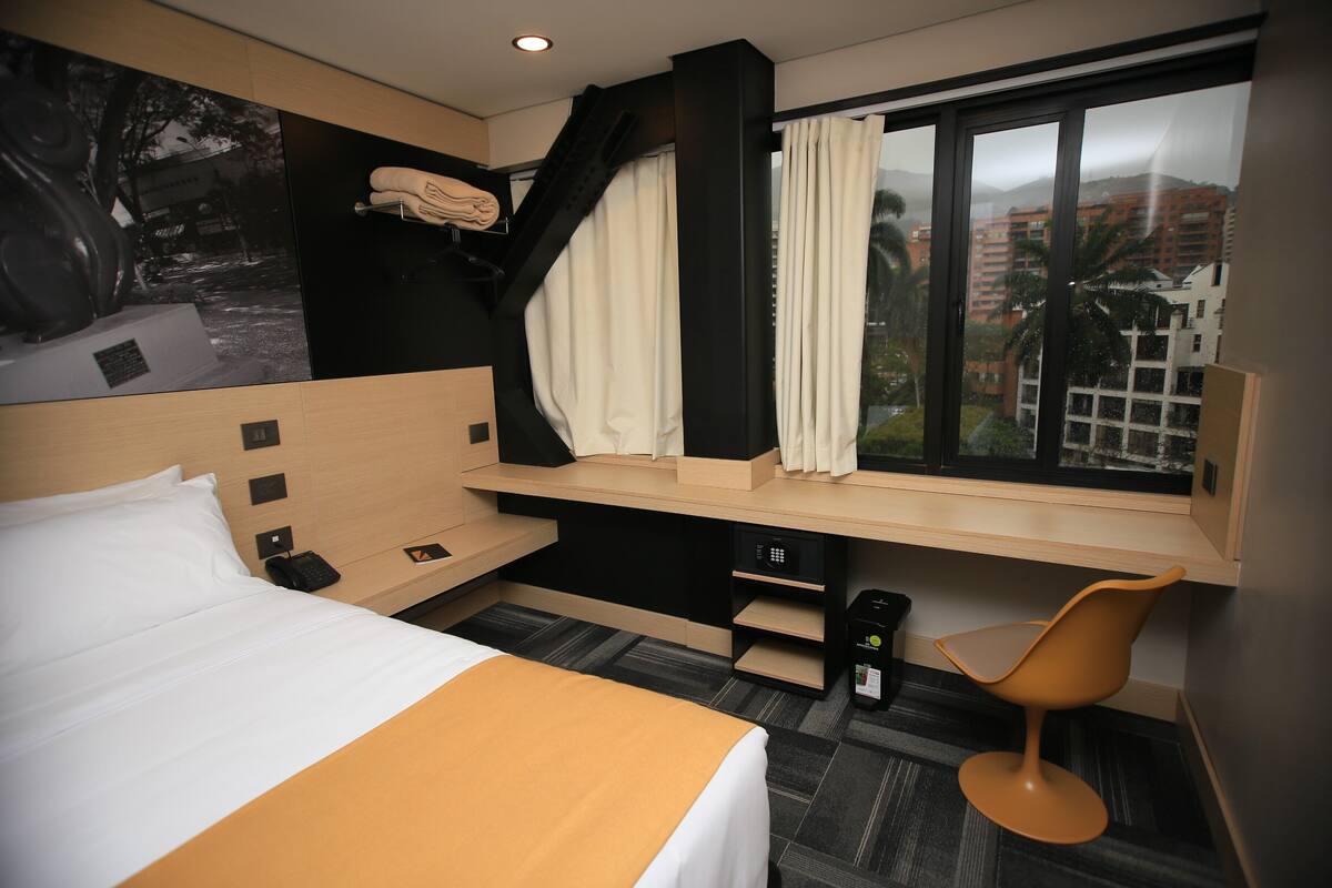 A hotel room with a bed, a desk, a chair, a window with curtains, and a wall-mounted shelf.