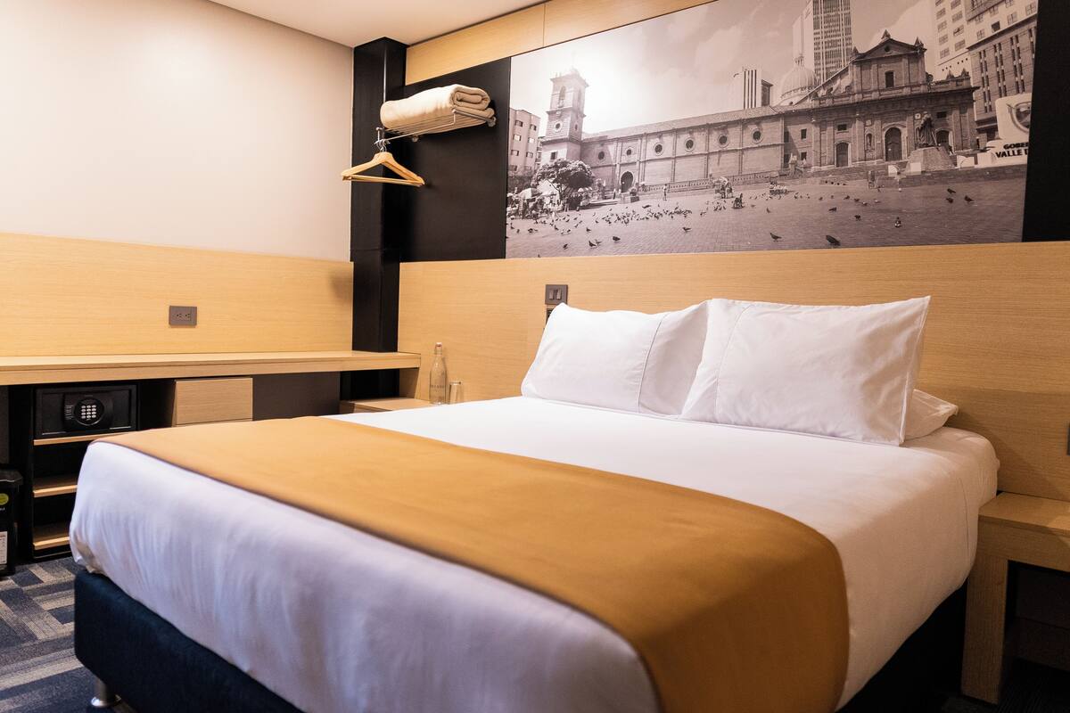 A hotel room with a large bed, a wooden headboard, a hanging towel, a small table, and a wall-mounted shelf.