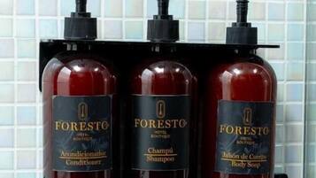 Un estante de baño con tres botellas de artículos de tocador FORESTO para hotel: acondicionador, champú y jabón corporal.