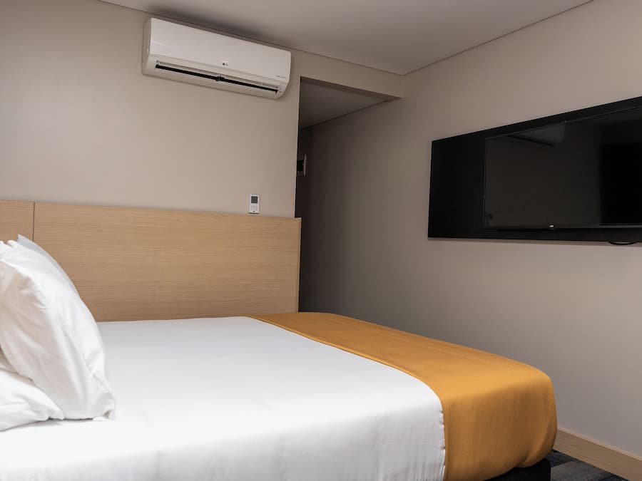 A hotel room with a bed, a television mounted on the wall, and an air conditioning unit above the bed.