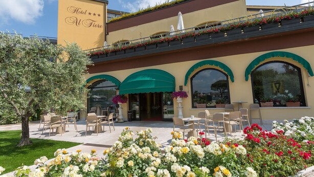 A hotel with a yellow facade, green awnings, outdoor seating, and flower beds. - Al Fiore (Peschiera del Garda)