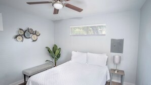 Un dormitorio con cama, ventilador de techo, banca, planta en maceta, mesita de noche con lámpara e interruptor de luz en la pared.