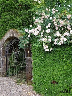 Une arche en pierre surmontée d’un portail en métal, entourée d’une végétation luxuriante et de roses roses en pleine floraison.
