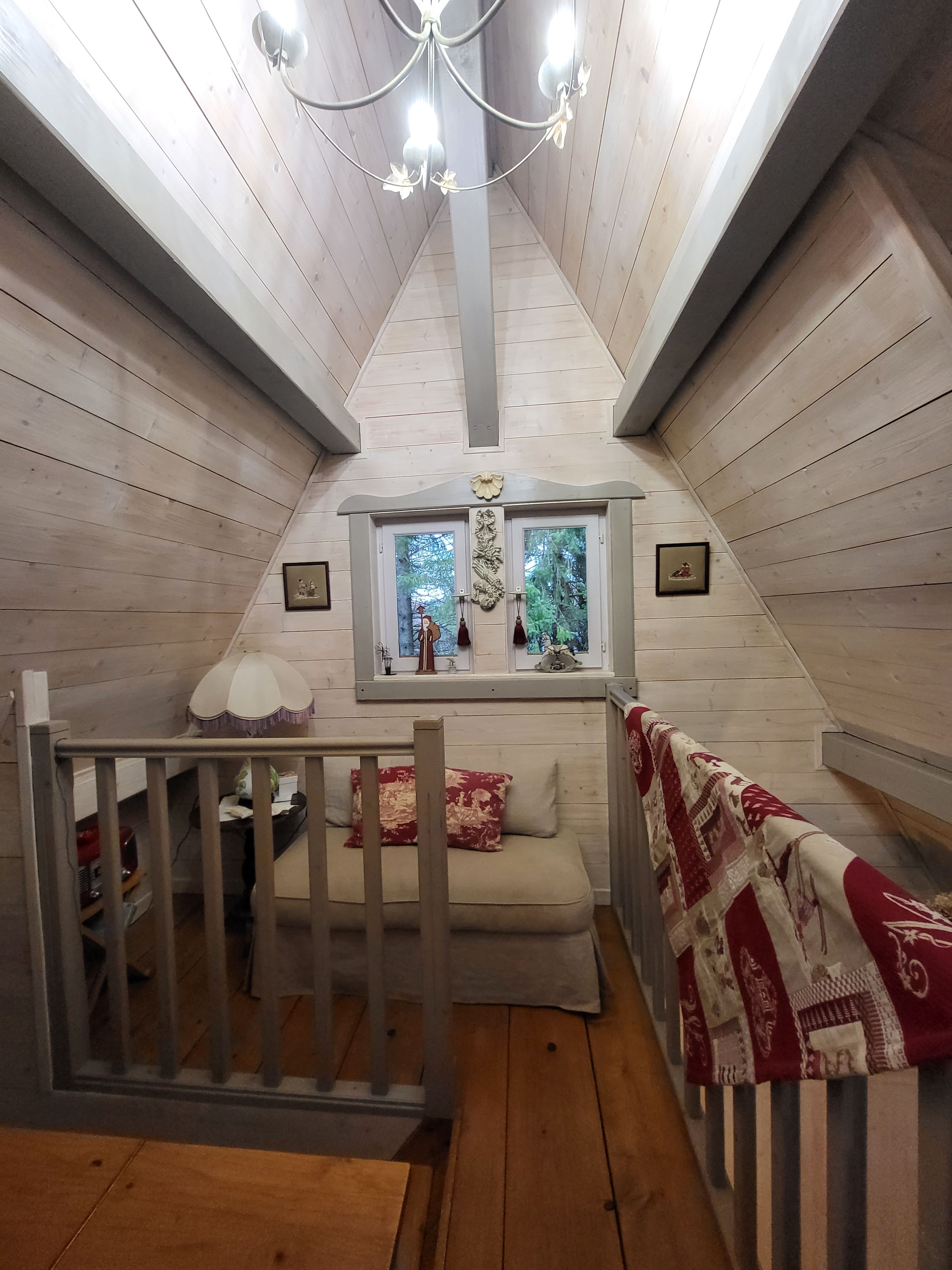 Una camera da letto accogliente in mansarda, con soffitto spiovente in legno, una finestra vista verde, un lampadario e un piccolo tavolo con una lampada.