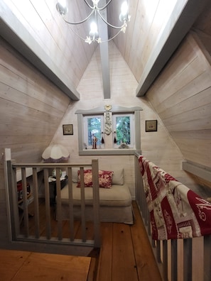 Ein gemütliches Schlafzimmer im Dachgeschoss mit holzgetäfeltem Giebeldach, einem Fenster mit Blick ins Grüne, einer Deckenleuchte und einem kleinen Tisch mit Lampe.