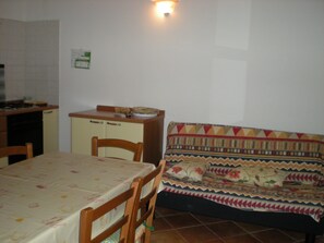 A compact kitchen with a dining table and chairs, a sofa with a patterned cover, and a wall-mounted light fixture.