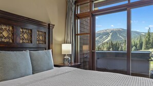 A hotel room with a large bed, a nightstand, a lamp, and a view of mountains through a large window. - Rare Ski In Ski Out Top Floor Corner Unit! (Sun Peaks)