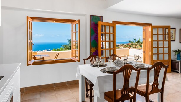 Un comedor con una mesa preparada para cuatro, ventanas grandes con marcos de madera y vista al mar.