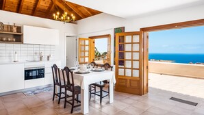 Un comedor con una mesa dispuesta para cuatro, sillas de madera, una araña y vistas al mar a través de puertas abiertas.