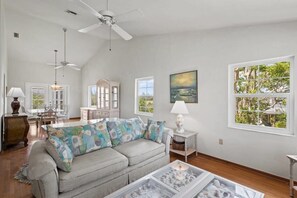 A spacious living room with a gray sofa, a glass coffee table, a ceiling fan, and a view of the outdoors through large windows.