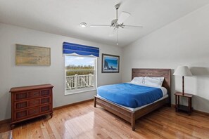 A bedroom with a bed, a dresser, a window with a view, a ceiling fan, and a lamp.