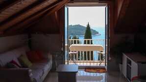 Ein gemütliches Zimmer mit Holzbalkendecke, einer Couch mit Kissen, einem kleinen Tisch und einem Balkon mit Tisch und Stühlen mit Blick auf eine malerische Wasserlandschaft und Hügel.
