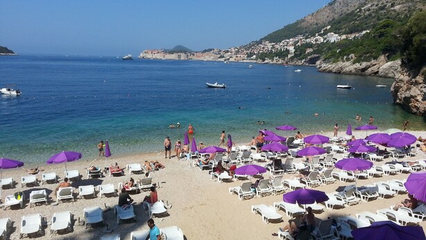 Ein Strand mit weißen Liegestühlen und violetten Sonnenschirmen, klarem, blauem Wasser und Booten in der Ferne.