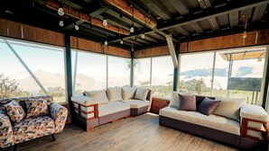A spacious outdoor seating area with a wooden floor, large windows offering a view of the landscape, and a ceiling with exposed beams and hanging lights. - Refugio Suncamal By Rotamundos (Cumanda)