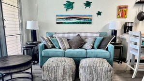 Ein senffarbenes Sofa mit vielen Kissen, zwei passende Hocker, ein Gemälde mit Strandmotiv und ein Schild mit der Aufschrift 'Surfing'.
