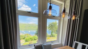 Comedor con mesa de madera, dos lámparas colgantes y vistas a un lago y colinas a través de amplios ventanales.