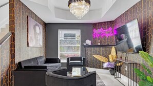 Een moderne woonkamer met een zwarte bank, een bijzettafeltje en een televisie op een standaard. Tegen de muur hangt een neonreclame en aan het plafond een kroonluchter.