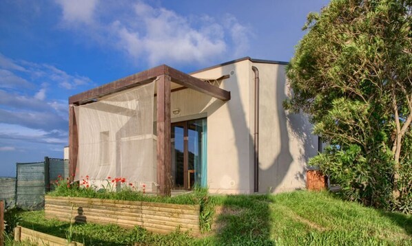 Une maison moderne dotée d’une grande fenêtre, d’une pergola en bois et d’un jardin agrémenté de fleurs rouges.