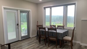 Ein Essbereich mit Tisch und Stühlen, große Fenster mit Blick auf Grünflächen und eine Glastür, die zu einem Außenbereich führt.