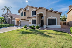 A two-story house with a beige exterior, a front garage, and a well-maintained lawn with small bushes.