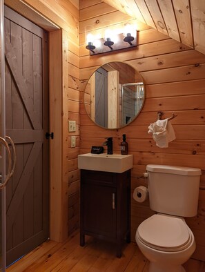 A wooden cabin bathroom with a wooden door, a round mirror, a sink with a soap dispenser, and a toilet. - La Bellota Cabin - Secluded, Hot Tub, Sauna, Game Room, Massage Chair (Laurelville)