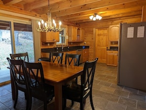 A wooden cabin interior with a dining table, chairs, a chandelier, a refrigerator, and wooden cabinets.