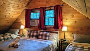 Un dormitorio cálido en el ático con una cama grande, dos camas más pequeñas, paredes de madera, una ventana con cortinas rojas y un ventilador de techo.