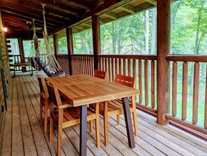 A wooden deck with a table and chairs, surrounded by glass windows offering a view of a forest. - La Bellota Cabin - Secluded, Hot Tub, Sauna, Game Room, Massage Chair (Laurelville)