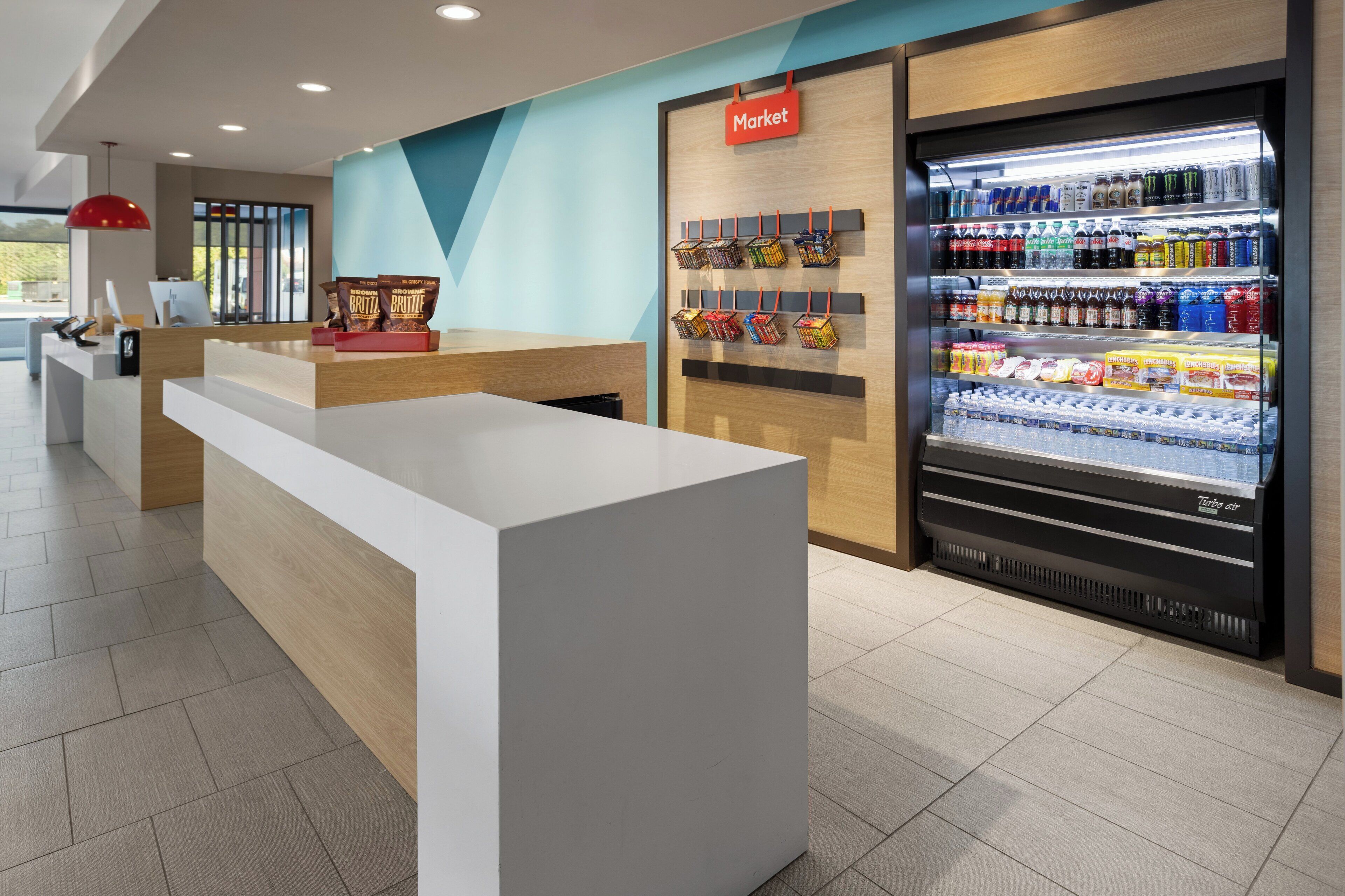 A modern reception area with a white reception desk, a 'Market' sign, and a refrigerated display case with various beverages.