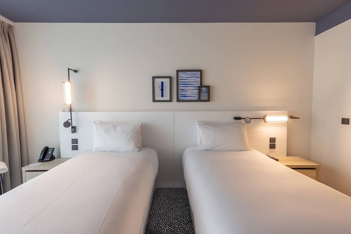 A hotel room with two single beds, a bedside table with a lamp, a phone, and a wall-mounted picture.