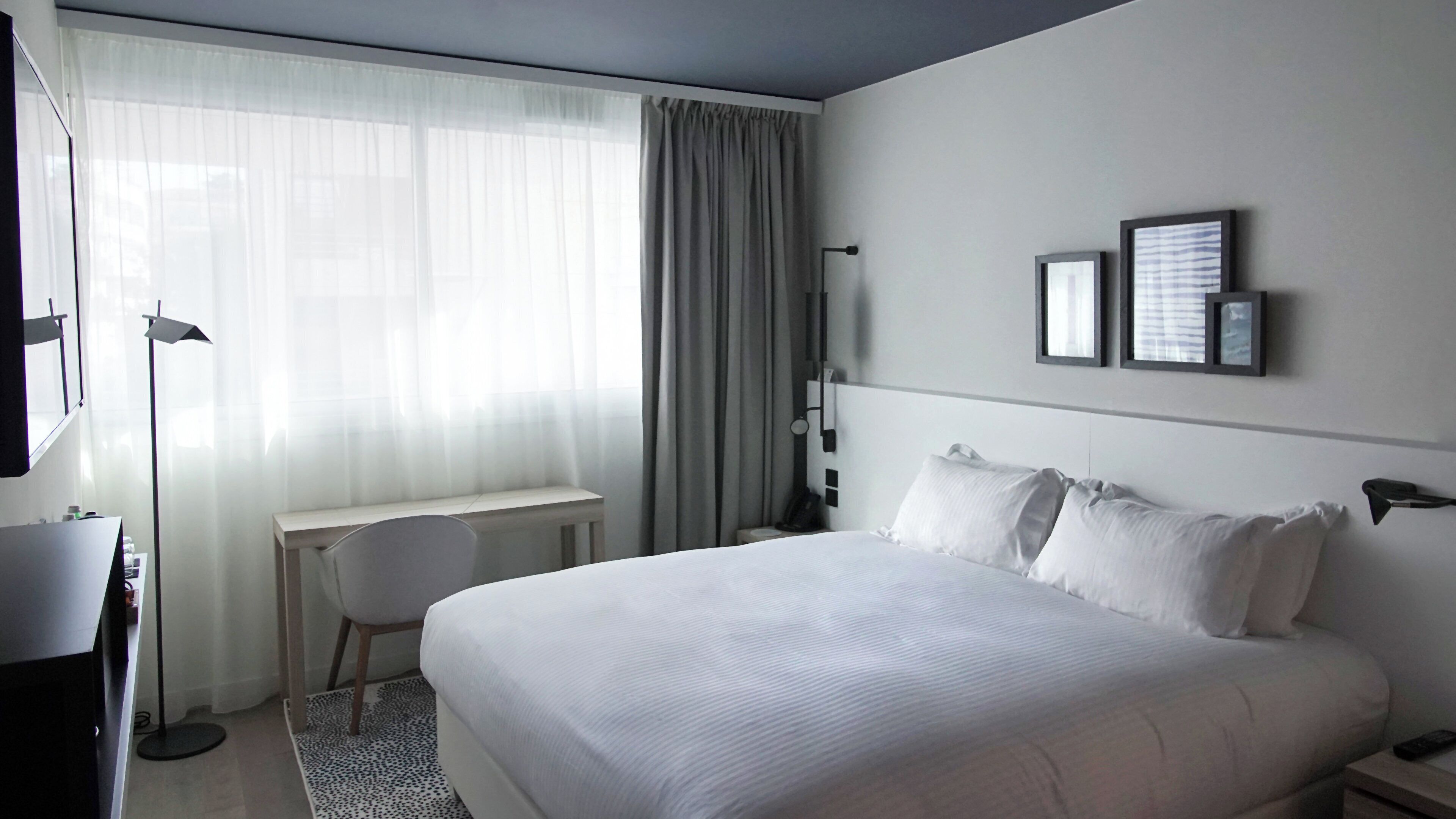A hotel room with a large bed, a desk, a chair, a desk lamp, and a window with curtains.