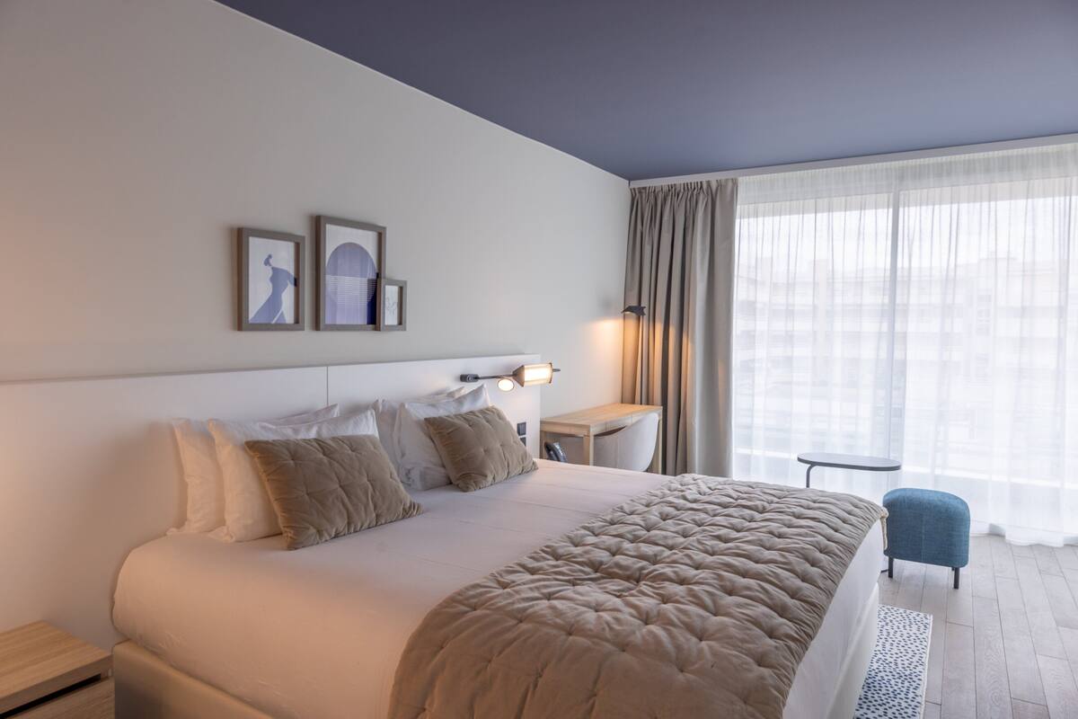 A modern hotel room with a large bed, a bedside table, a chair, and a window with curtains.
