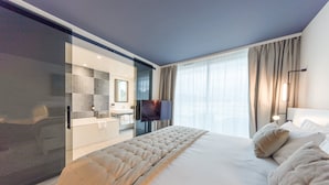 A modern hotel room with a large bed, a television, a bathroom with a glass shower enclosure, and a large window with curtains. - Hotel Indigo Cagnes-sur-Mer by IHG (Cagnes-sur-Mer)