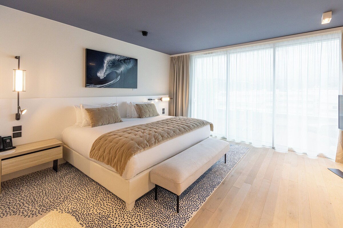 A modern hotel room with a large bed, a flat-screen TV, a bedside table with a lamp, and a large window with sheer curtains.