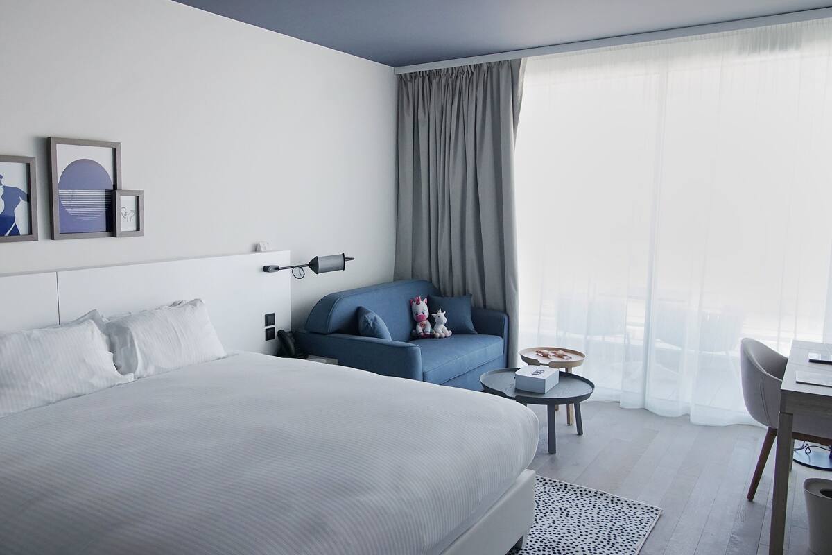 A modern hotel room with a large bed, a blue armchair, a small round table, and a view of the city through sheer curtains.