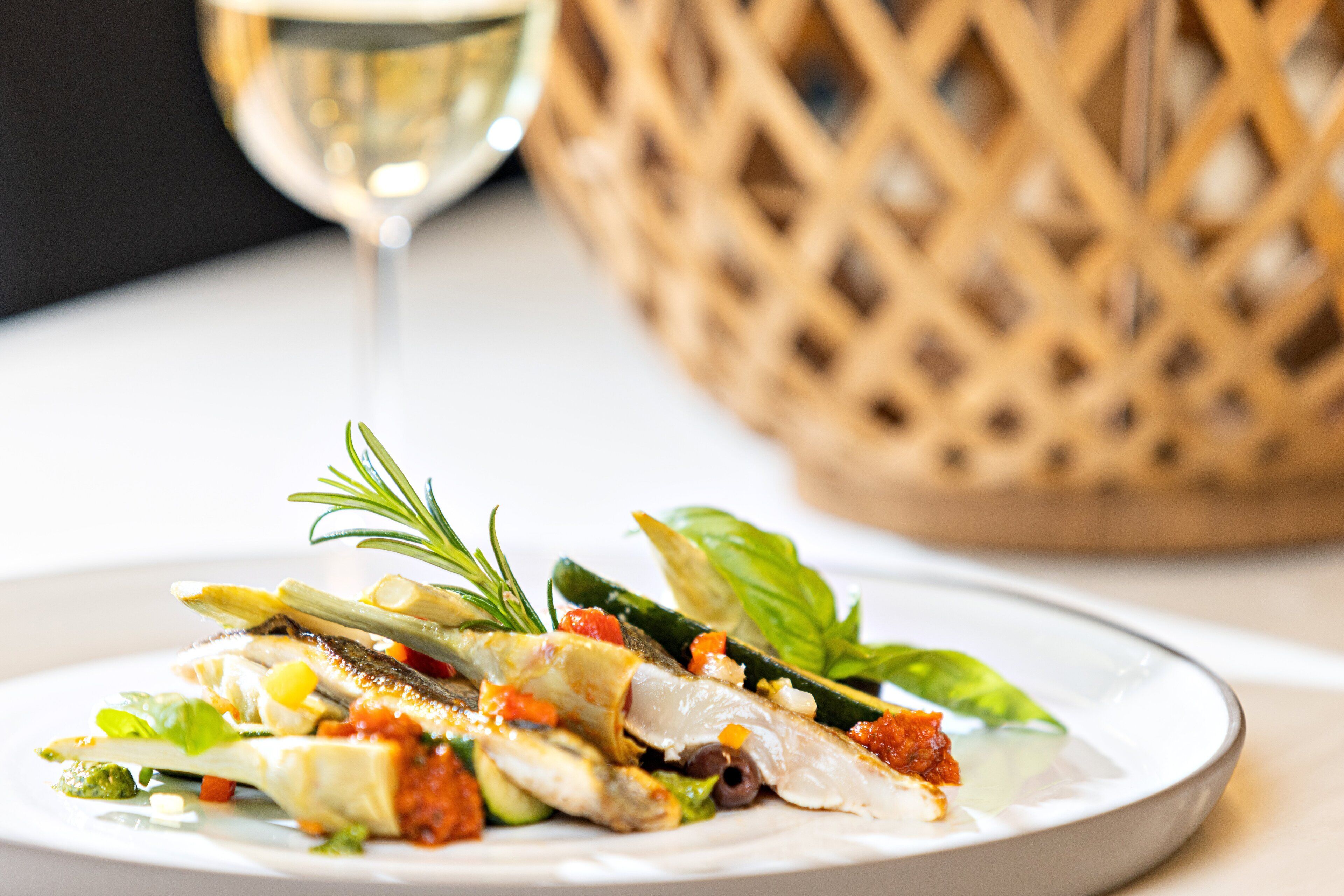 A plate of food with various vegetables and herbs, a glass of white wine, and a woven basket in the background.