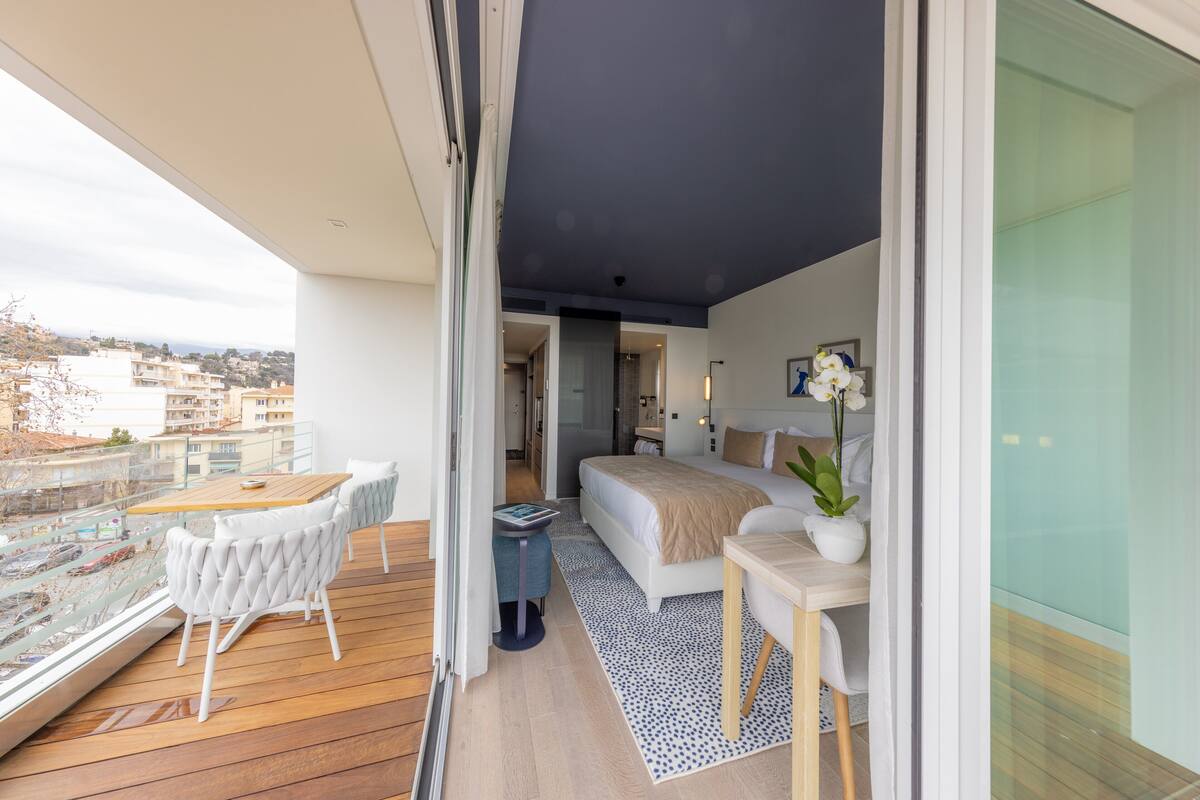 A modern bedroom with a large bed, a small table, and a chair. There is a balcony with a wooden floor and white chairs.