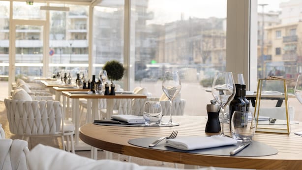 A restaurant with white chairs and tables, some with wine glasses and bottles, and a view of buildings through large windows. - Hotel Indigo Cagnes-sur-Mer by IHG (Cagnes-sur-Mer)