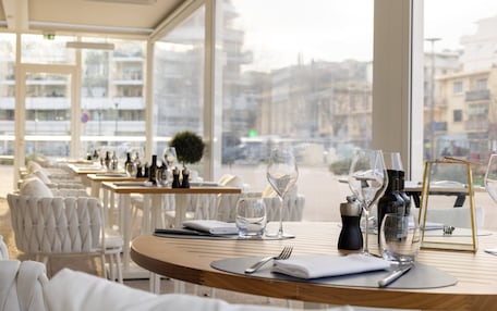A restaurant with white chairs and tables, some with wine glasses and bottles, and a view of buildings through large windows.